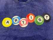 A close-up of a blue mineral-wash t-shirt featuring a graphic spelling 'ARTIST' using colorful circular pool balls in yellow, blue, pink, orange, green, and black.