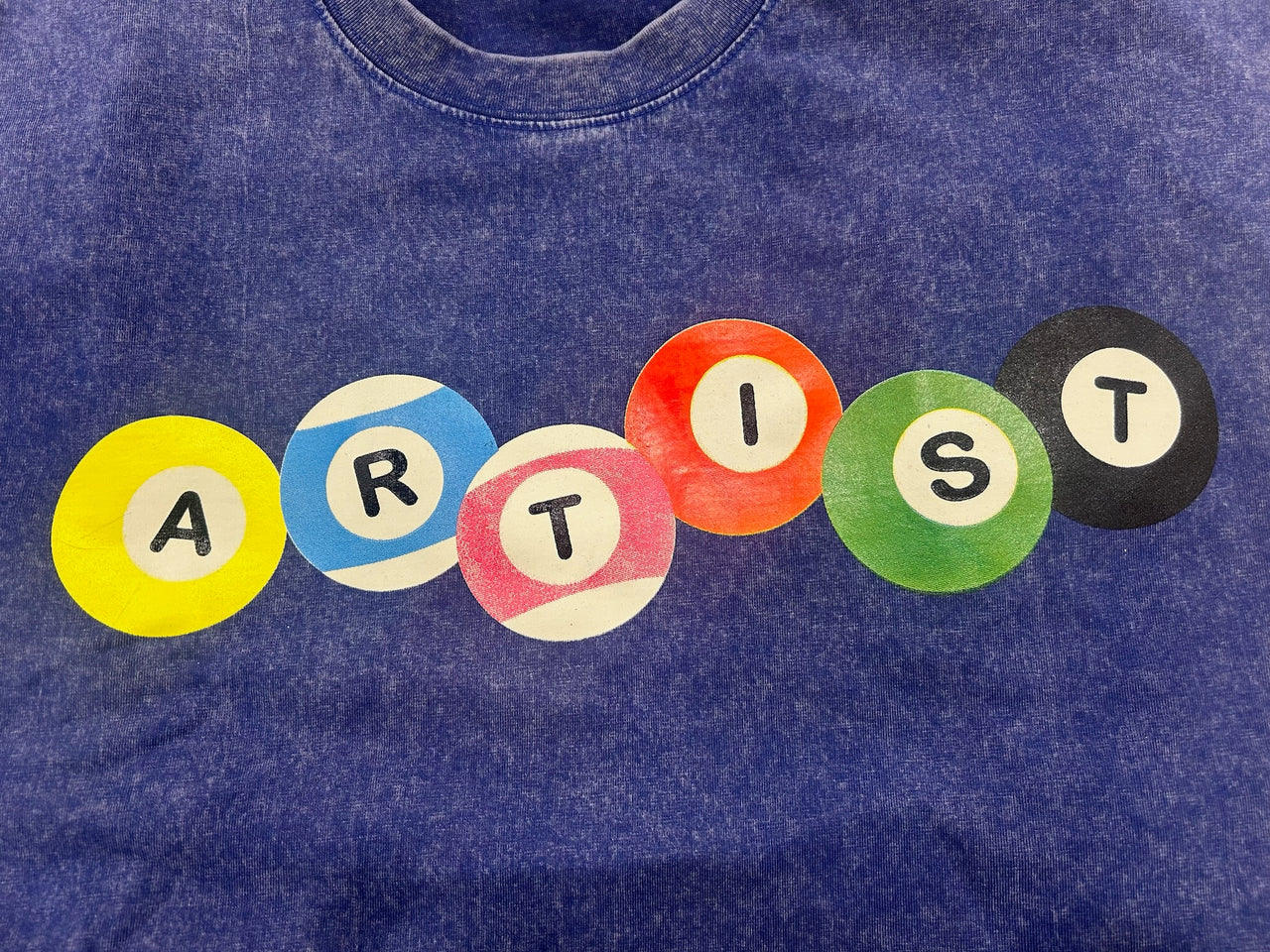 A close-up of a blue mineral-wash t-shirt featuring a graphic spelling 'ARTIST' using colorful circular pool balls in yellow, blue, pink, orange, green, and black.