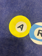A close-up of a blue textured fabric surface featuring two circular letter patches. The left patch is bright yellow with a white center and a black letter 'A', while the right patch is light blue and white with a black letter 'R'.