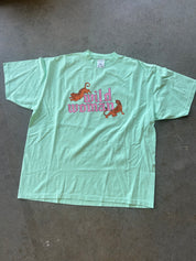 A light mint green short-sleeve t-shirt laid flat on a gray concrete floor. The front features a graphic with the words 'wild woman' in pink script, flanked by two orange wildcats