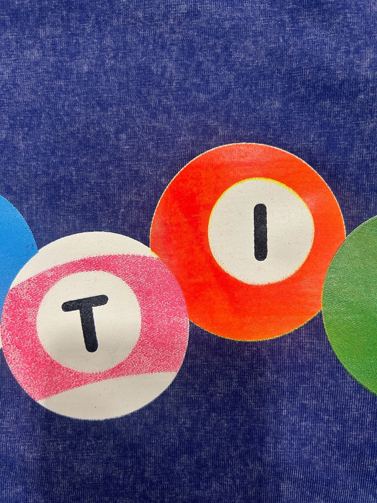 A close-up of a blue textured fabric surface with three overlapping circular painted patches: a blue patch with a black T, an orange patch with a black I, and a green patch partially visible on the right.