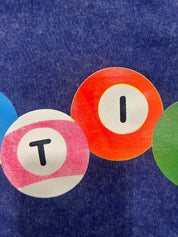 A close-up of a blue textured fabric surface with three overlapping circular painted patches: a blue patch with a black T, an orange patch with a black I, and a green patch partially visible on the right.