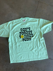 A light mint green short-sleeve t-shirt with a black graphic text reading 'DON'T FORGET TO HAVE FUN!' and a small yellow smiley face, laid flat on a gray concrete floor.