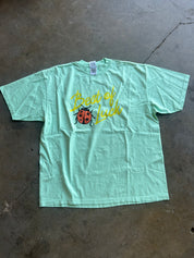 A mint green short-sleeve t-shirt laid flat on a concrete floor, featuring a graphic print of a red ladybug and the yellow text 'Great Luck'.