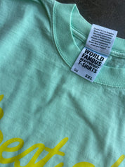 Close-up of a mint green t-shirt showing the inner neck label which reads 'WORLD FAMOUS HEAVYWEIGHT T-SHIRTS' and '3XL', with a hint of yellow script text on the chest.