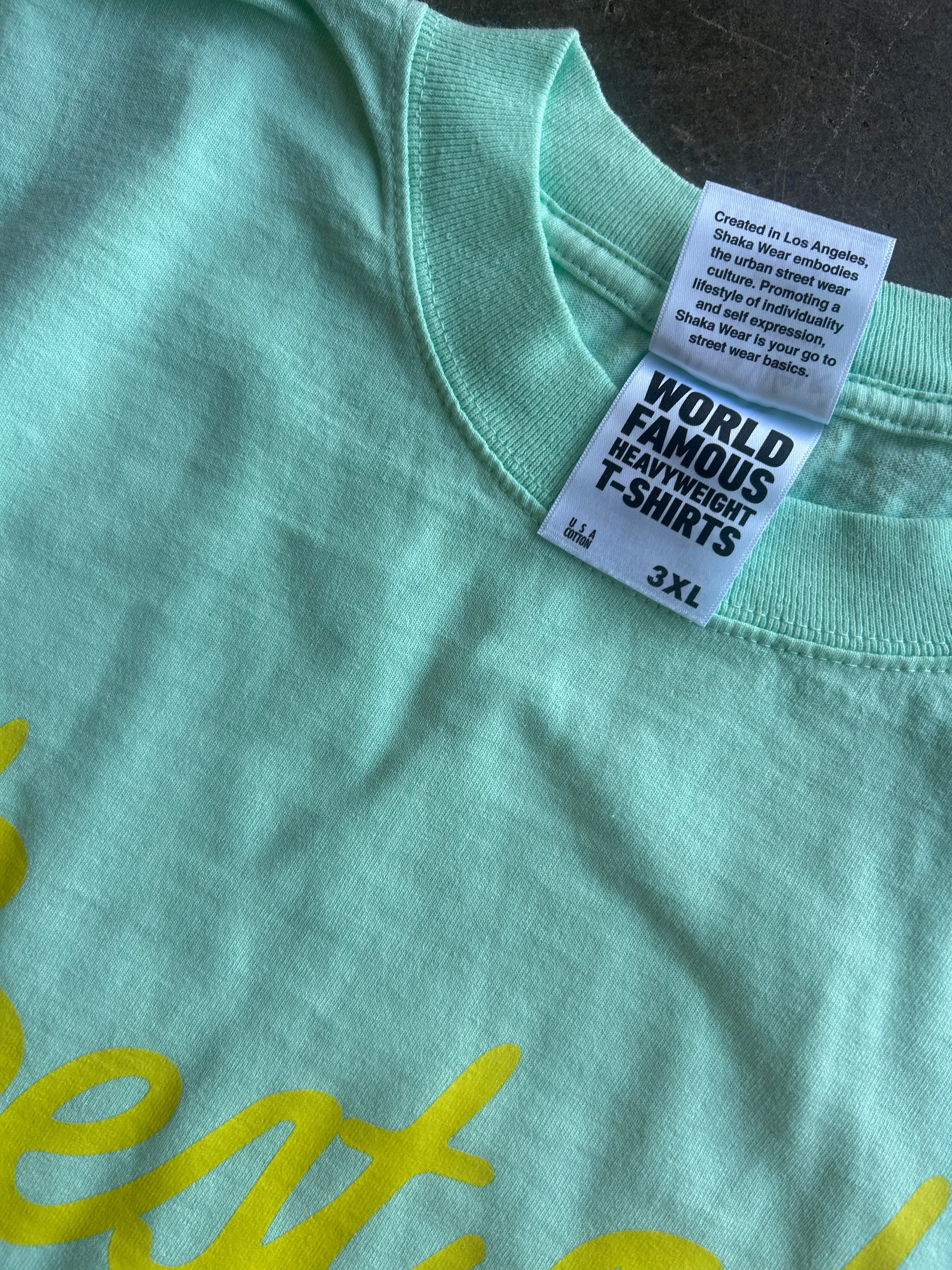 Close-up of a mint green t-shirt showing the inner neck label which reads 'WORLD FAMOUS HEAVYWEIGHT T-SHIRTS' and '3XL', with a hint of yellow script text on the chest.
