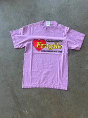 A light purple short-sleeve t-shirt laid flat on a gray concrete floor, featuring a graphic print of a red heart with the word 'Fragile' in yellow and black text reading 'I'm emotionally fragile please handle with care'.