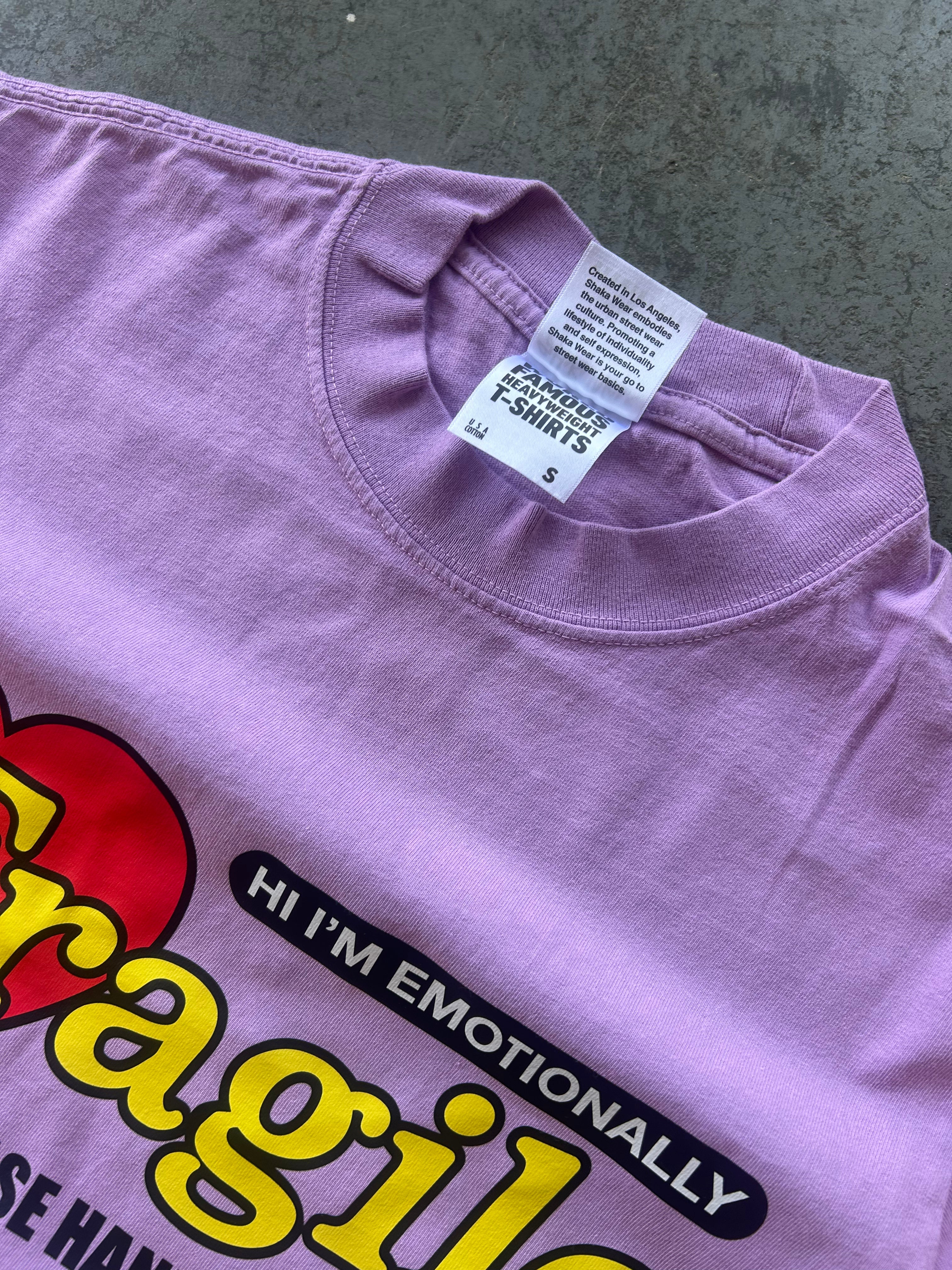 A close-up of a lavender purple t-shirt showing the inner collar with a white Shaka brand tag and part of a colorful graphic print featuring yellow text and a red heart.