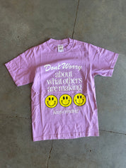 Purple graphic tee with the text Don't Worry about what others are making 3 smiley faces Keep Creating on a concrete surface