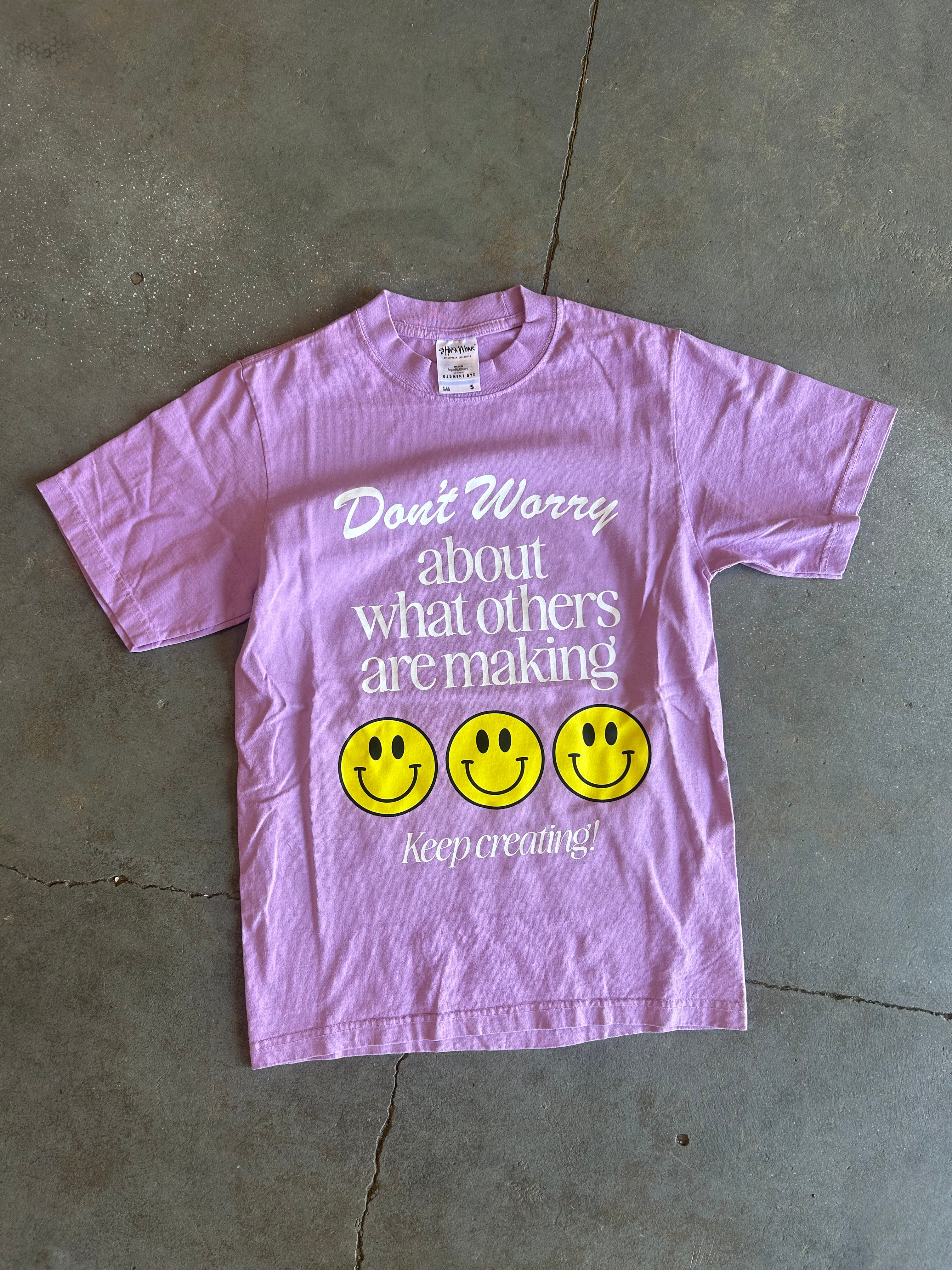 Purple graphic tee with the text Don't Worry about what others are making 3 smiley faces Keep Creating on a concrete surface