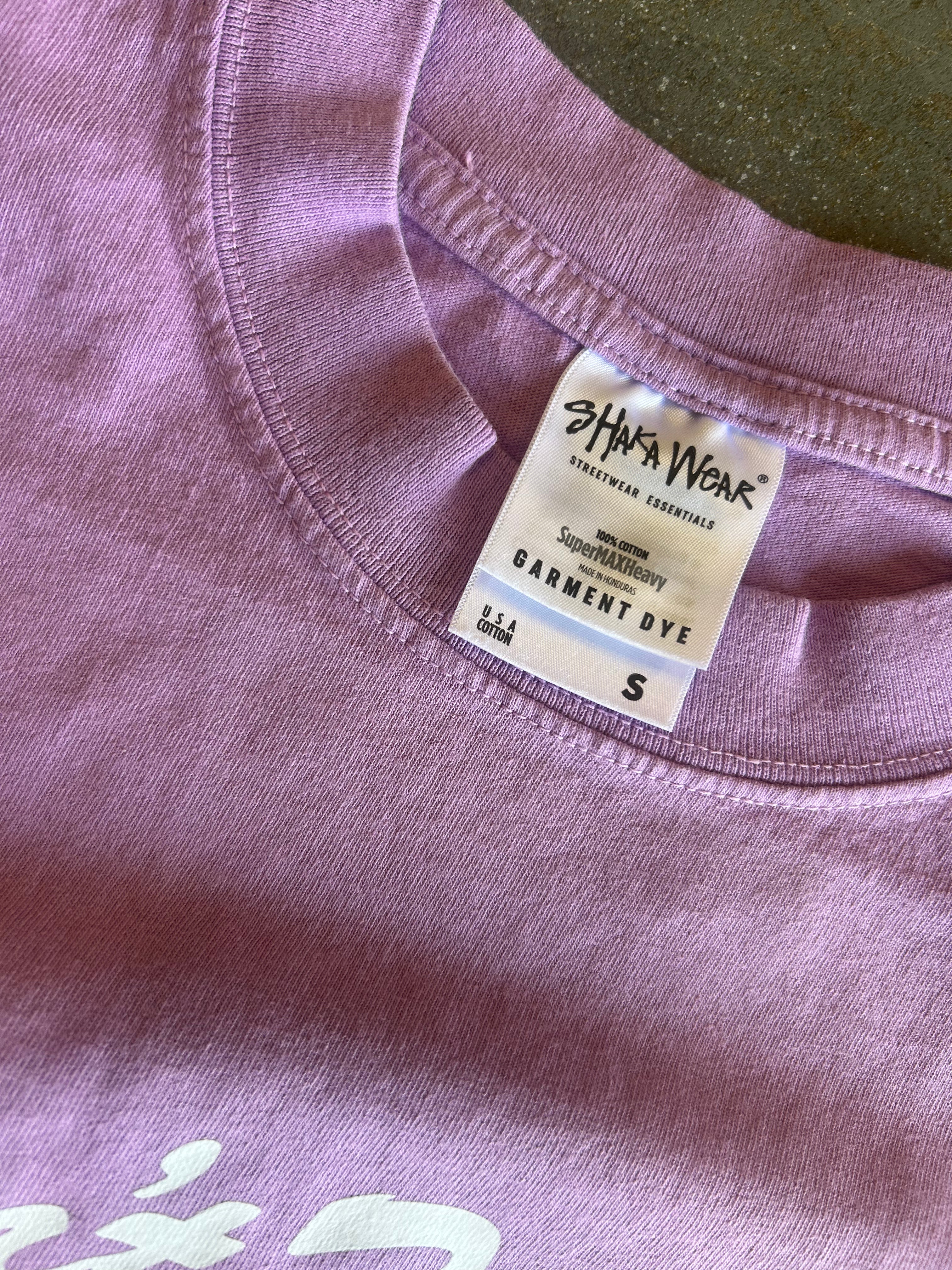 Close-up of a lavender-purple t-shirt showing the inner collar with a white 'Shaka Wear' garment dye tag.