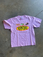 A light purple short-sleeve t-shirt laid flat on a gray concrete floor, featuring a large 'Burntoast' oval logo in yellow and red on the front, along with smaller sponsor-style graphics above it.