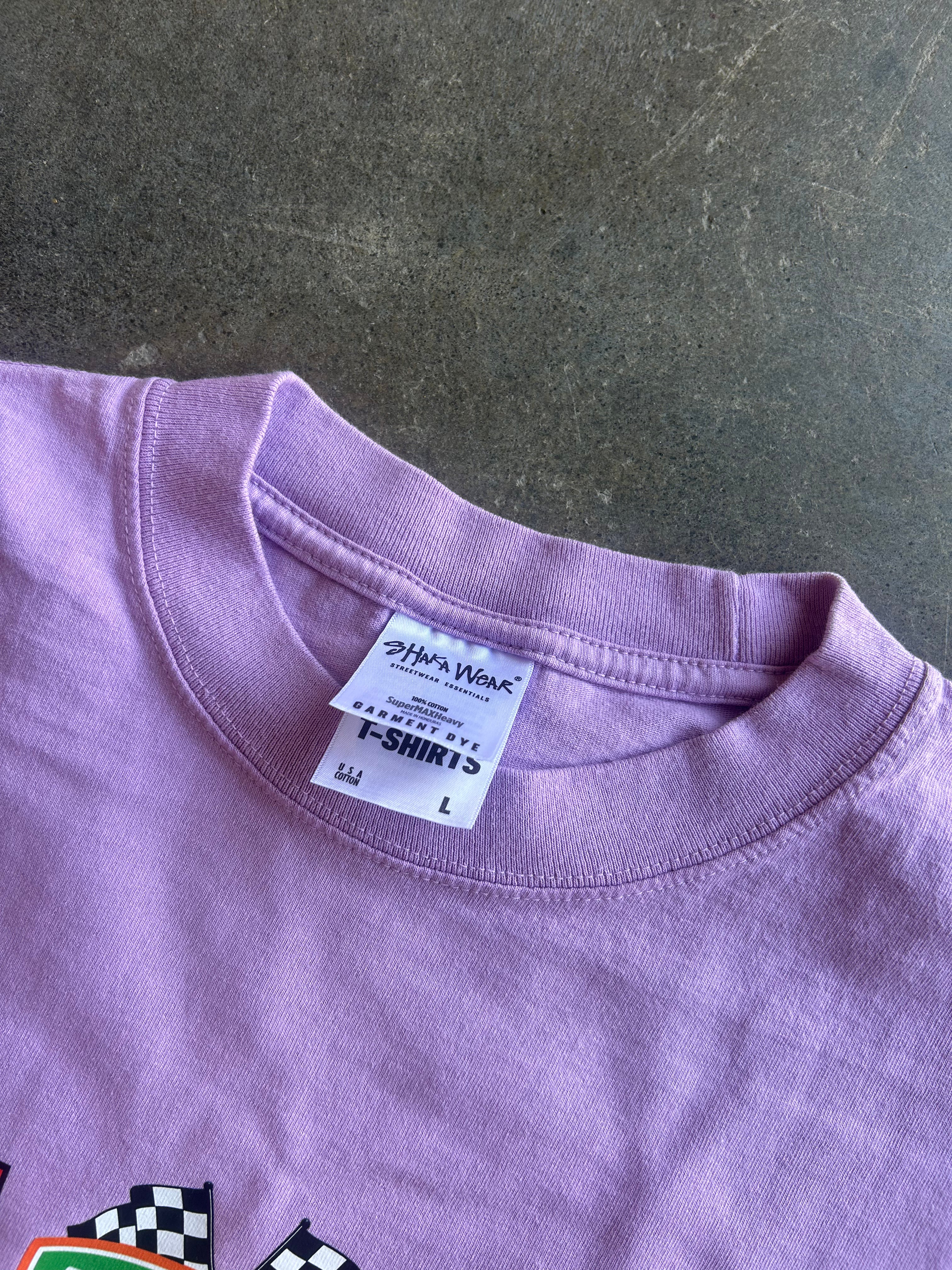 A close-up of a lavender purple t-shirt showing the inner collar label which reads 'Other Wear' and 'T-SHIRTS', laid flat on a dark textured surface.