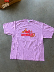 A light purple short-sleeve t-shirt with a red graphic reading 'wild woman' and two orange tigers, laid flat on a gray concrete floor.