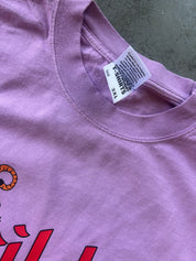 Close-up of a lavender-purple t-shirt showing the inner neck label which reads 'T-SHIRTS' and '3XL', with a small red graphic and orange circular detail visible at the bottom left.