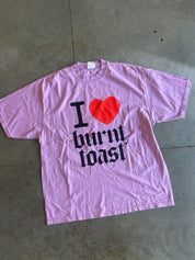 A light pink short-sleeve t-shirt with a graphic print reading 'I [red heart] burnt toast' in black gothic-style lettering, laid flat on a gray concrete floor.