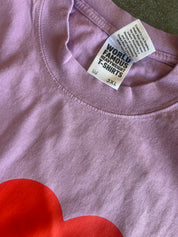 Close-up of a lavender-purple t-shirt with a red heart graphic at the bottom and a white neck label that reads 'WORLD FAMOUS HEARTED T-SHIRTS'.