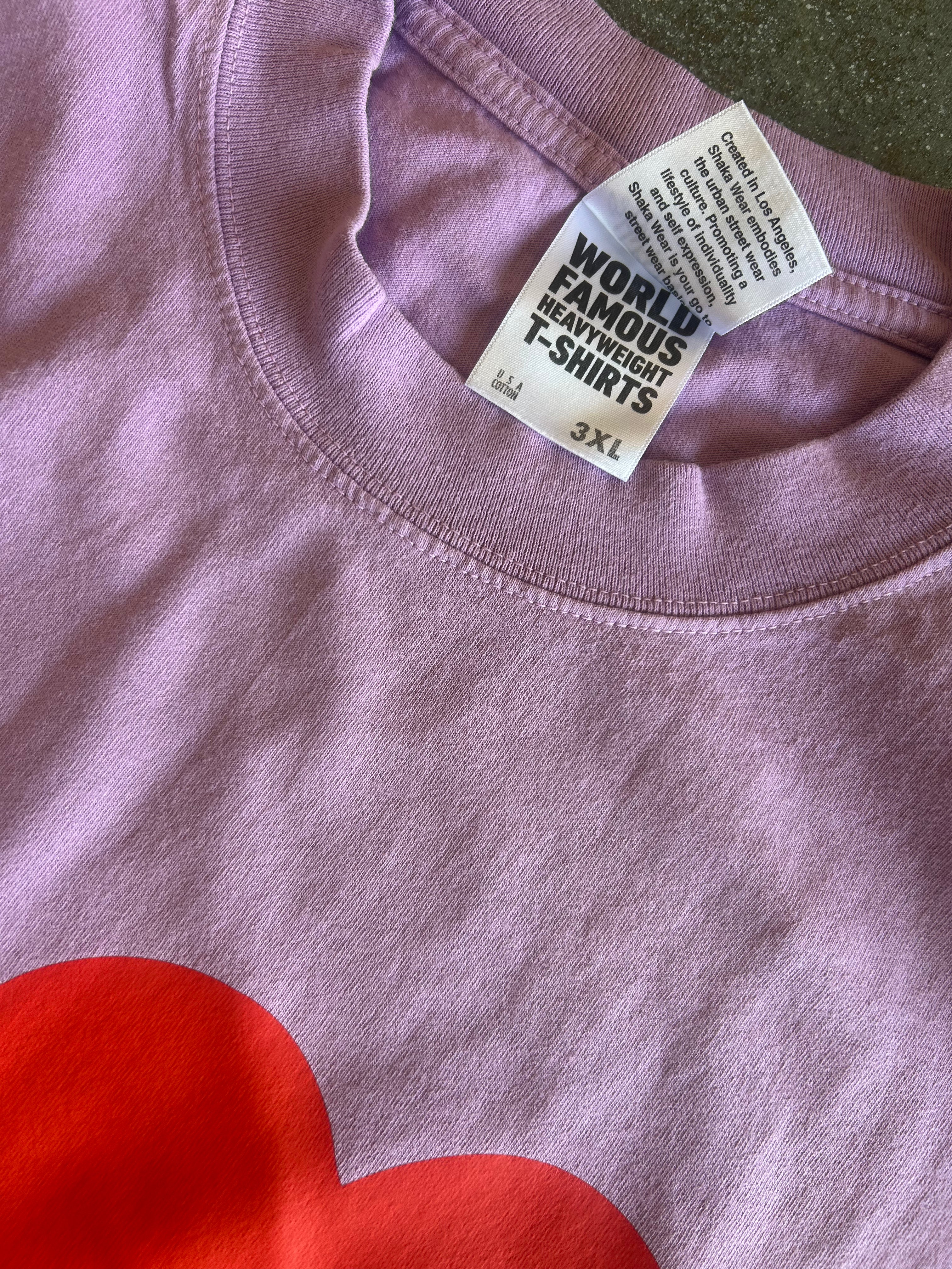 Close-up of a lavender-purple t-shirt with a red heart graphic at the bottom and a white neck label that reads 'WORLD FAMOUS HEARTED T-SHIRTS'.