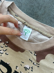 Close-up of a beige and brown printed t-shirt with a hand pointing to the inner neck label which reads 'COTTON UNLIMITED' and 'XXXLARGE'.