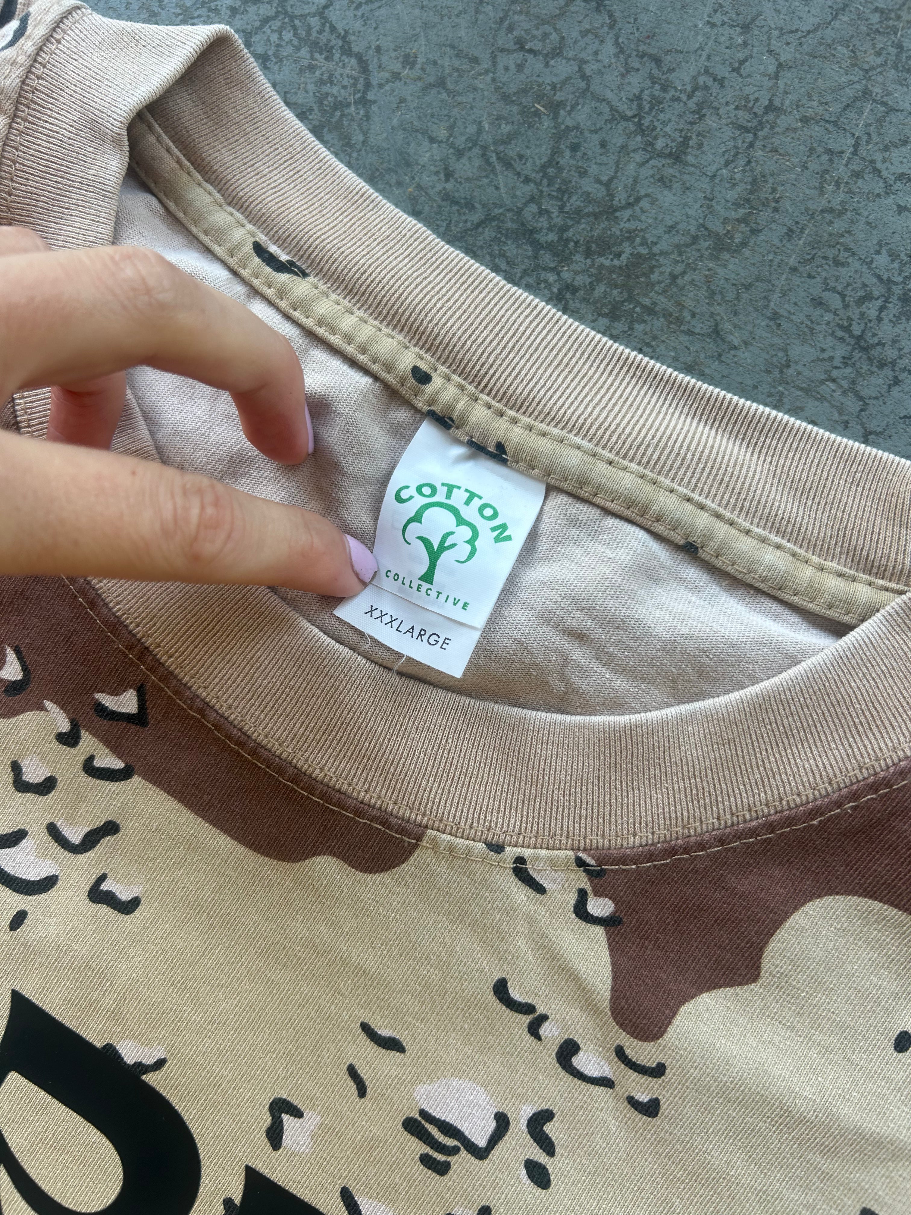 Close-up of a beige and brown printed t-shirt with a hand pointing to the inner neck label which reads 'COTTON UNLIMITED' and 'XXXLARGE'.