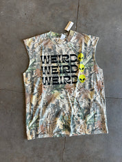 A sleeveless camouflage graphic shirt laid flat on a gray concrete floor. The shirt features a woodland camo pattern in shades of green, brown, and beige, with the word 'WEIRD' printed three times in black block letters across the chest, and three yellow alien heads aligned vertically on the right side.