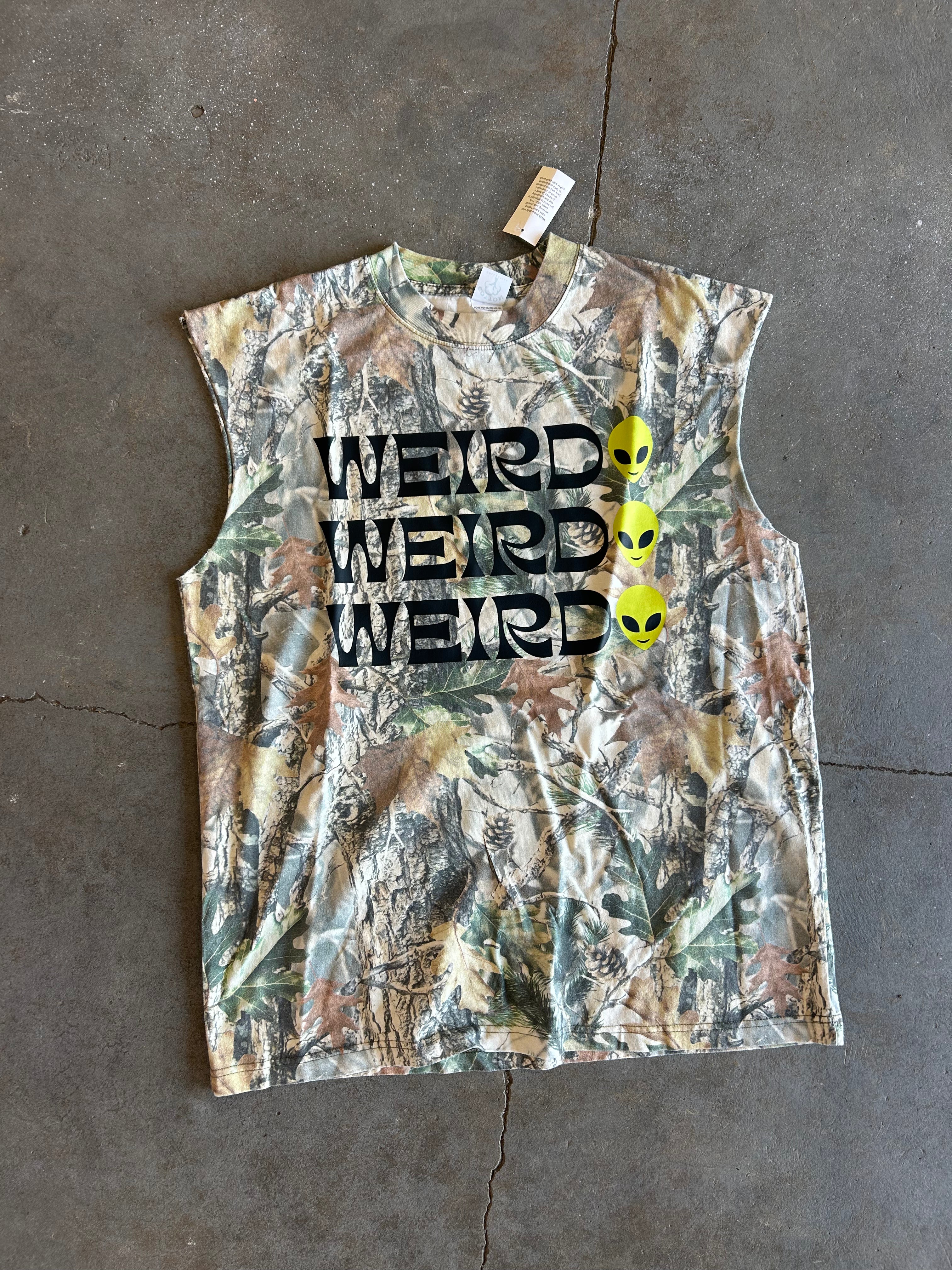 A sleeveless camouflage graphic shirt laid flat on a gray concrete floor. The shirt features a woodland camo pattern in shades of green, brown, and beige, with the word 'WEIRD' printed three times in black block letters across the chest, and three yellow alien heads aligned vertically on the right side.