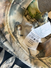 Close-up of a camouflage-patterned garment showing the inner collar and a sewn-in care label. The label reads 'Buckeye' and 'LARGE', with a woodland camo print in beige, brown, green, and gray tones.