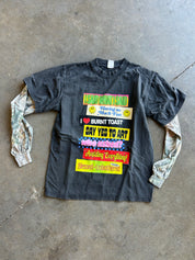 A black long-sleeve graphic t-shirt with a colorful text print on the front, laid flat on a concrete floor. The sleeves are made from camouflage-patterned fabric.