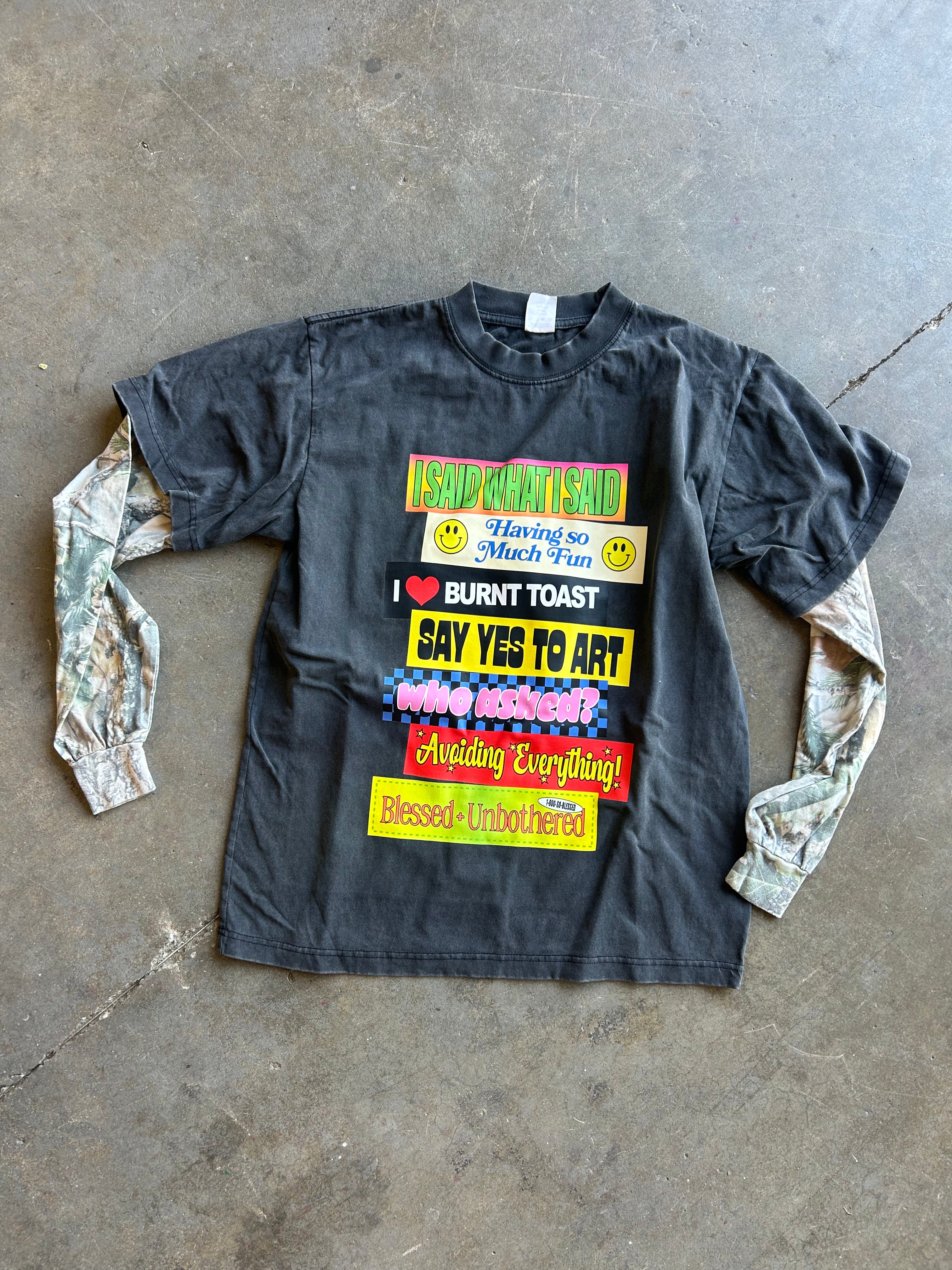 A black long-sleeve graphic t-shirt with a colorful text print on the front, laid flat on a concrete floor. The sleeves are made from camouflage-patterned fabric.