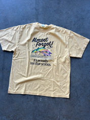 Yellow graphic tee with tex that reads 'Almost forget its not that serious" with a dolphin on a concrete surface