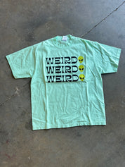 Seafoam green graphic tee with weird alien x3 on a concrete surface
