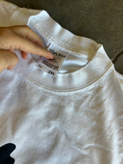 A close-up of a white crewneck t-shirt laid flat on a dark textured surface, with a hand pointing to the inner neck label which reads 'CHINA MADE' and '2XL'.