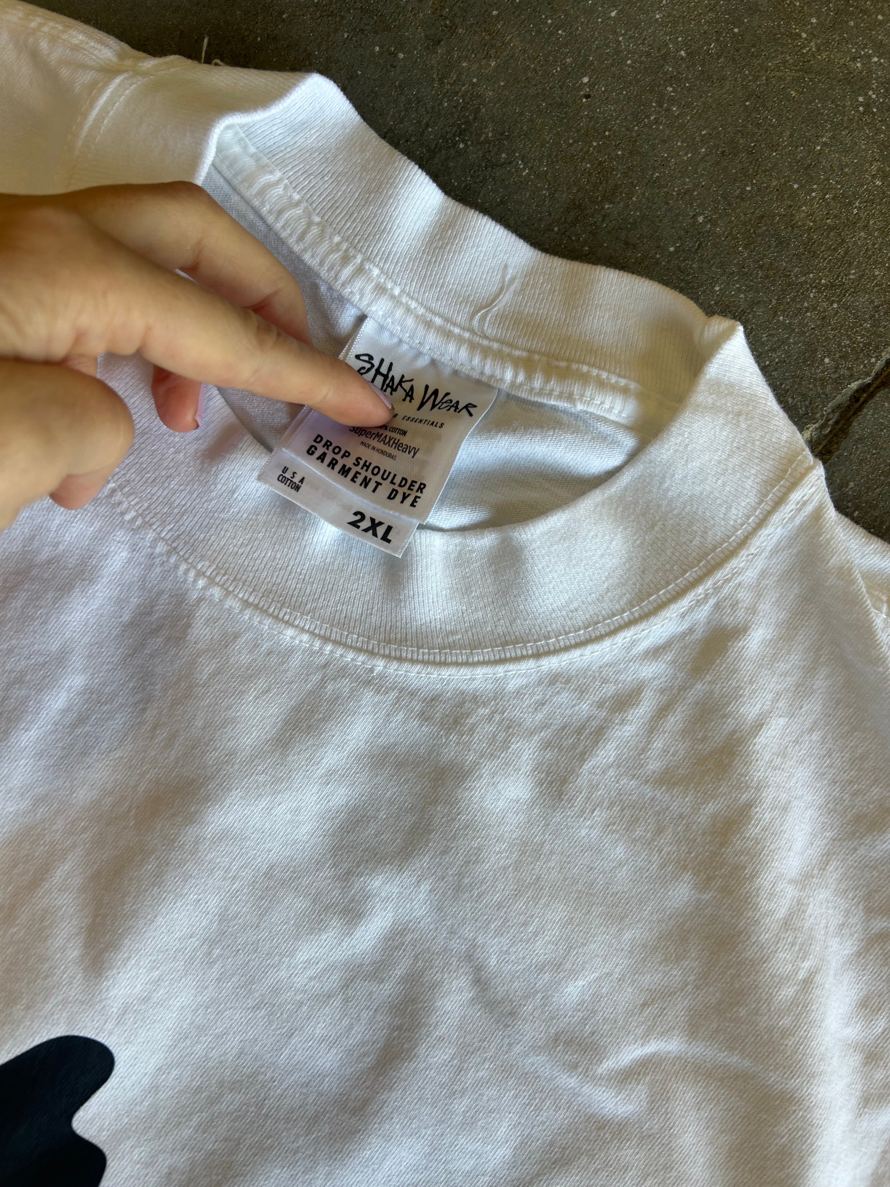 A close-up of a white crewneck t-shirt laid flat on a dark textured surface, with a hand pointing to the inner neck label which reads 'CHINA MADE' and '2XL'.