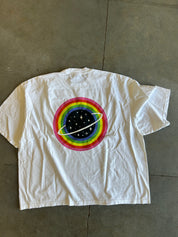 A white short-sleeve t-shirt laid flat on a gray concrete floor, featuring a large circular graphic on the back with concentric rainbow rings, a black starry center, and a white ring around it.