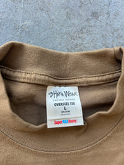 Close-up of the inside collar of a tan oversized cotton tee, showing the brand label 'Shaka Wear' and 'Super MAX Heavy' text on a gray concrete background.