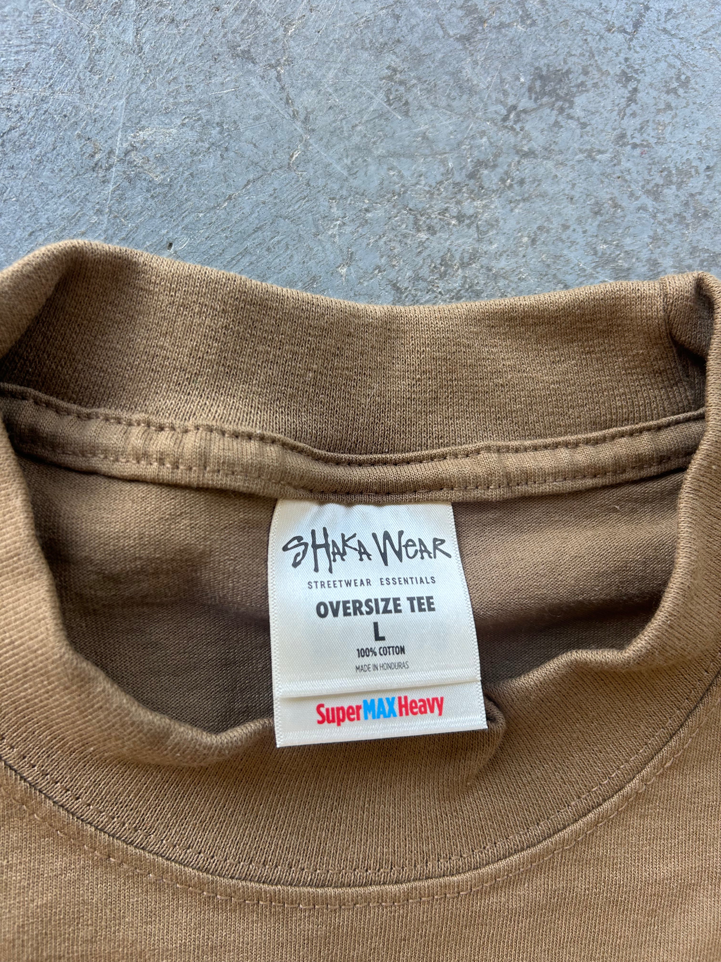 Close-up of the inside collar of a tan oversized cotton tee, showing the brand label 'Shaka Wear' and 'Super MAX Heavy' text on a gray concrete background.