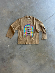 A tan long-sleeve t-shirt with a colorful graphic on the chest, laid flat on a gray concrete floor. The graphic features a rainbow arch with the text 'Actually it is all rainbows and butterflies'.