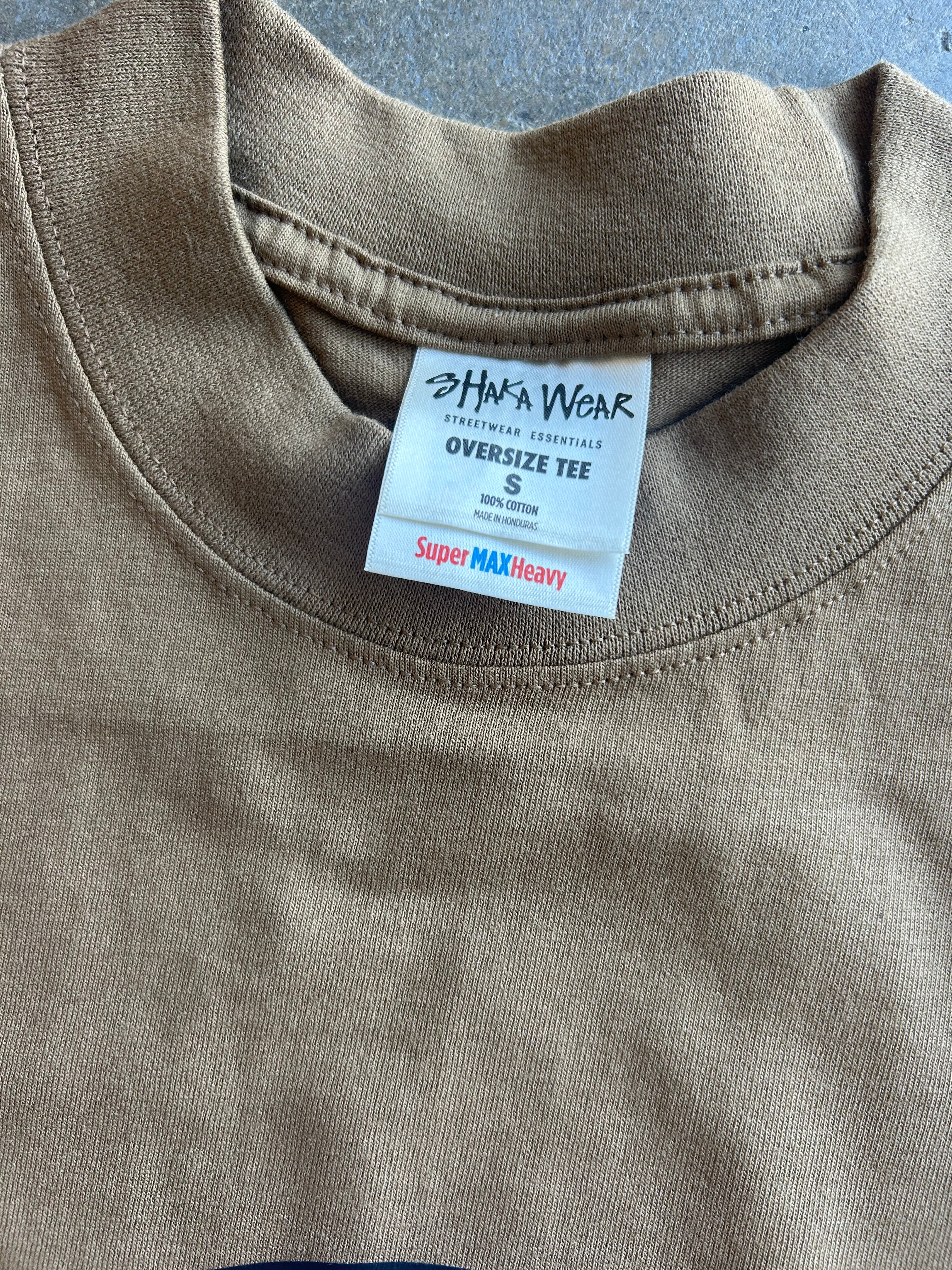 Close-up of an oversized crewneck t-shirt in a muted taupe-brown color, showing the inner neck label for 'Sha'a Wear'.