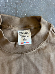 A close-up of a taupe brown oversized crewneck t-shirt laid flat on a gray concrete surface, showing the inner neck label for 'Shaka Wear'.