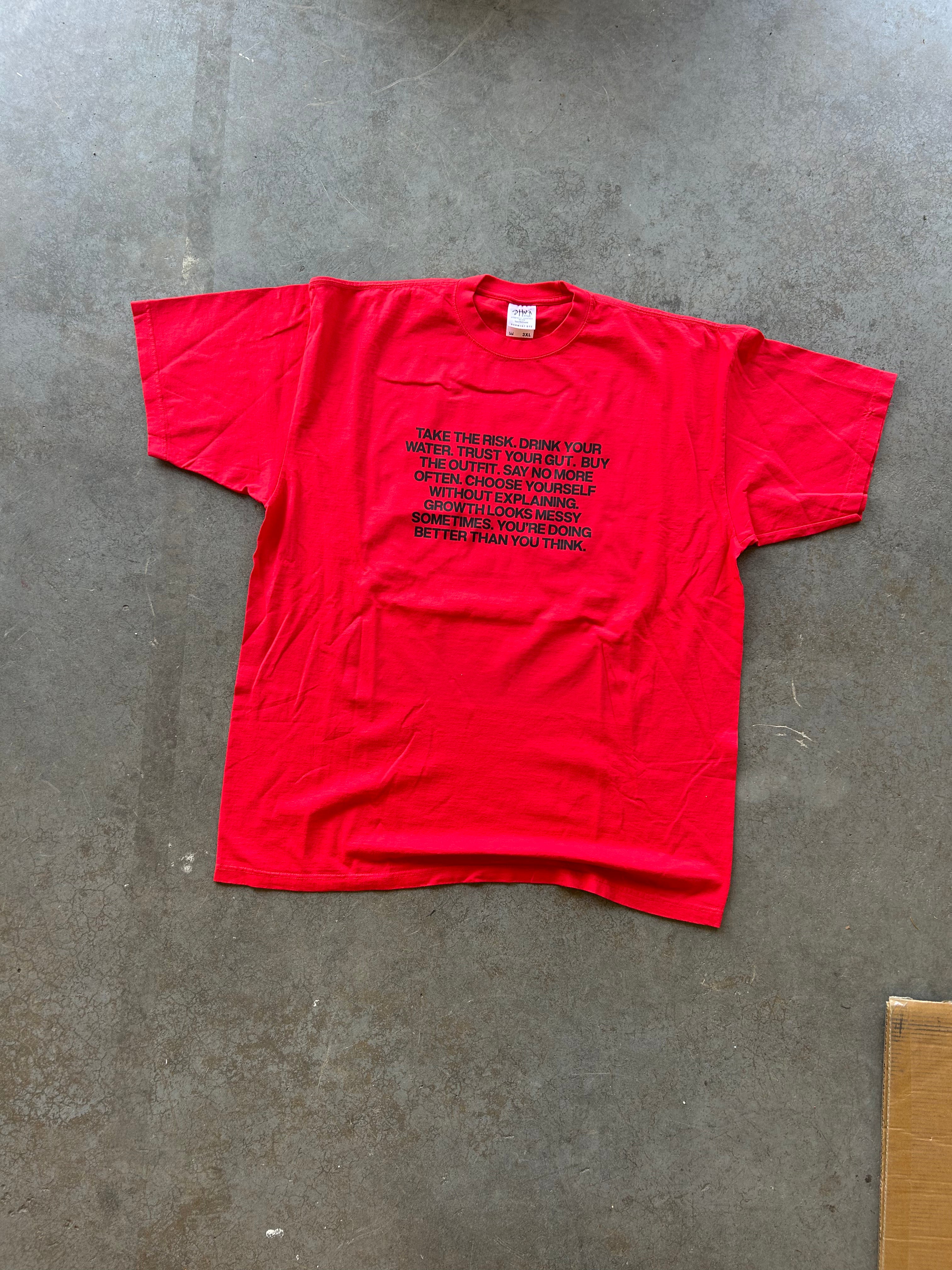 A bright red short-sleeve t-shirt with black printed text on the front, laid flat on a gray concrete floor.