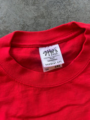 A close-up of a bright red crewneck t-shirt showing the inner neck label which reads 'SHAKA Streetwear Essentials' and 'Garment Dye 3XL'. The t-shirt is laid flat on a textured gray concrete surface.