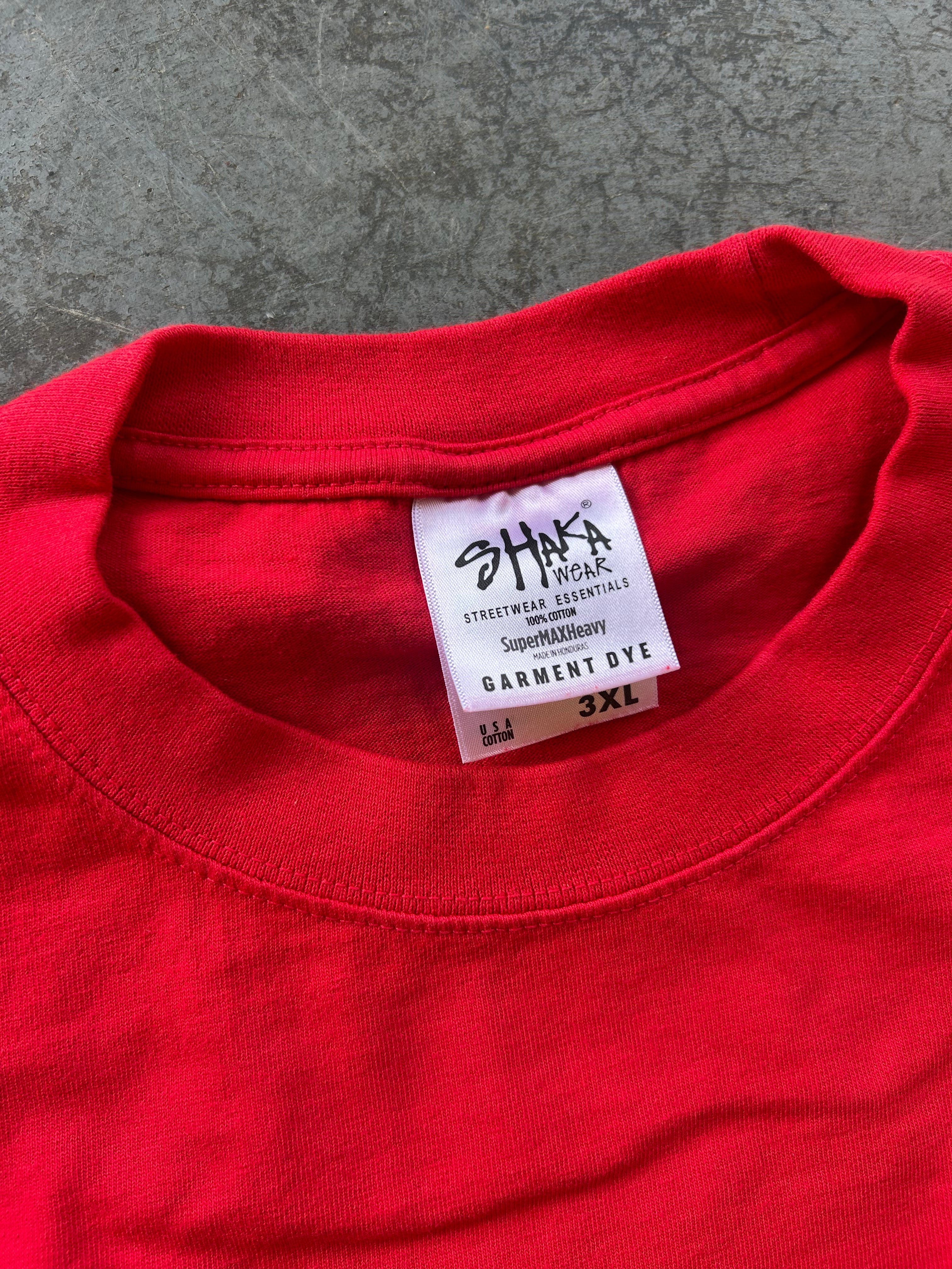 A close-up of a bright red crewneck t-shirt showing the inner neck label which reads 'SHAKA Streetwear Essentials' and 'Garment Dye 3XL'. The t-shirt is laid flat on a textured gray concrete surface.