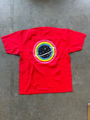 A bright red short-sleeve t-shirt laid flat on a gray concrete floor, featuring a retro circular graphic on the chest with concentric rainbow rings and a black planet with white stars.