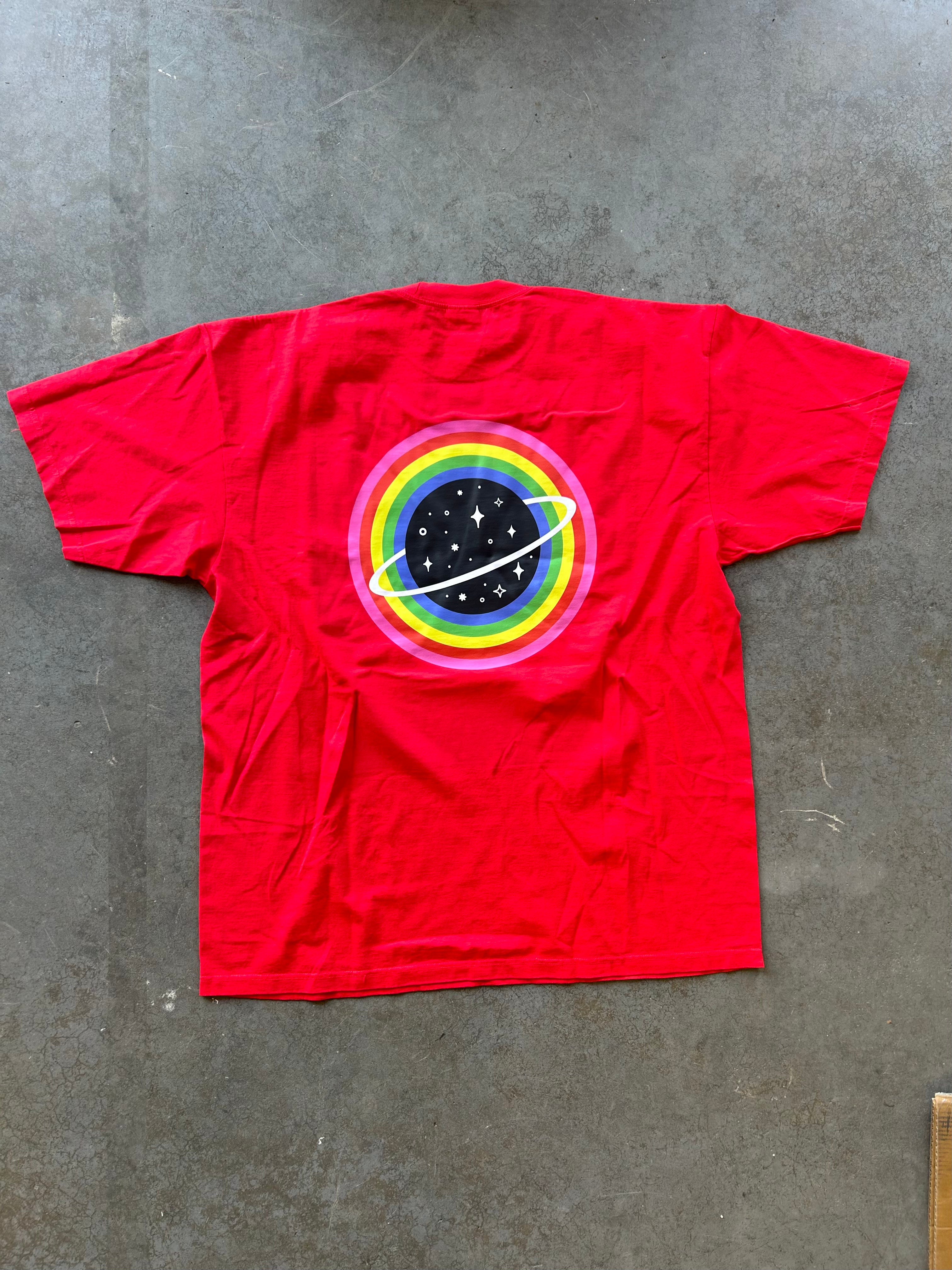 A bright red short-sleeve t-shirt laid flat on a gray concrete floor, featuring a retro circular graphic on the chest with concentric rainbow rings and a black planet with white stars.