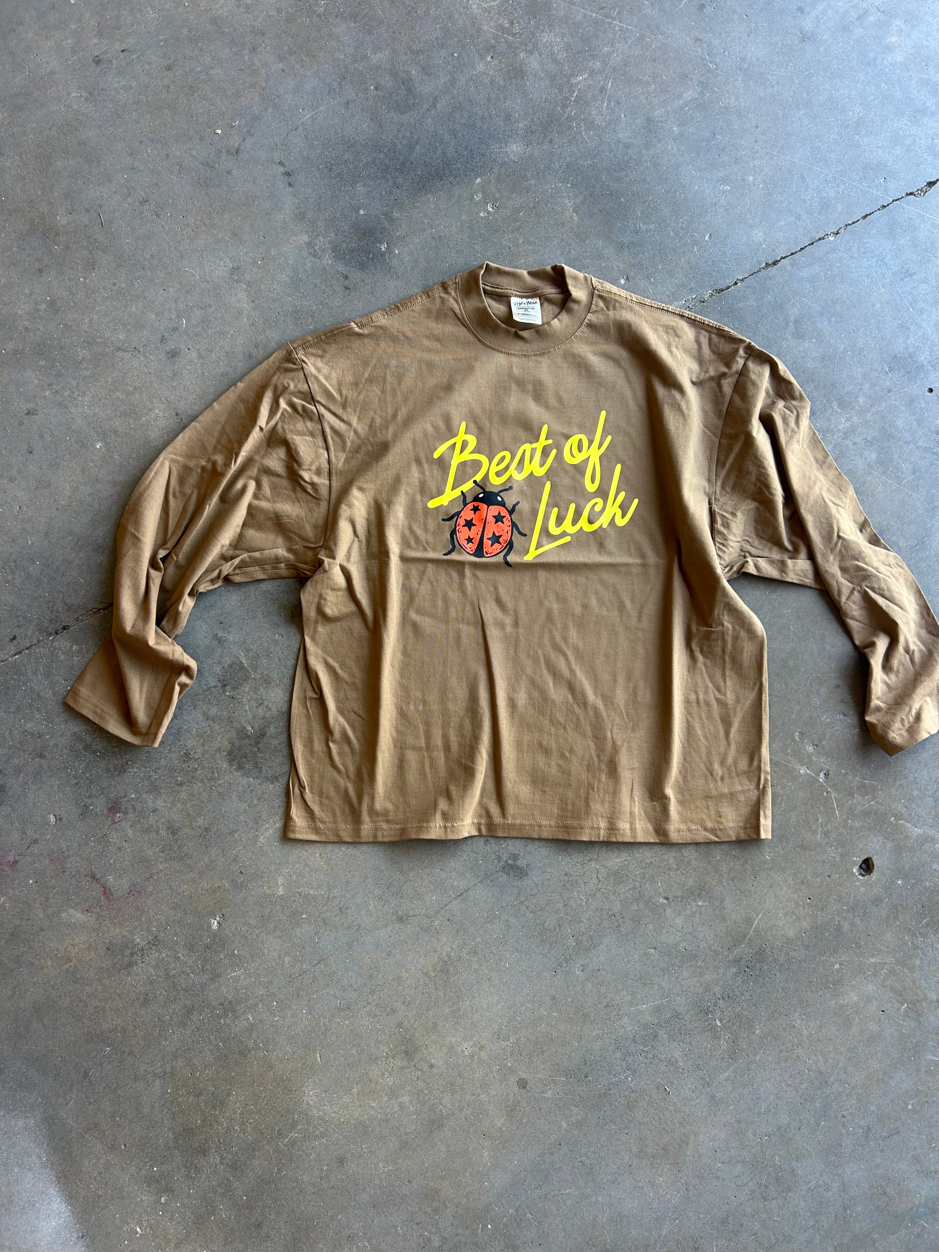A tan long-sleeve t-shirt with a 'Beat of Luck' graphic featuring a ladybug, laid flat on a gray concrete floor.