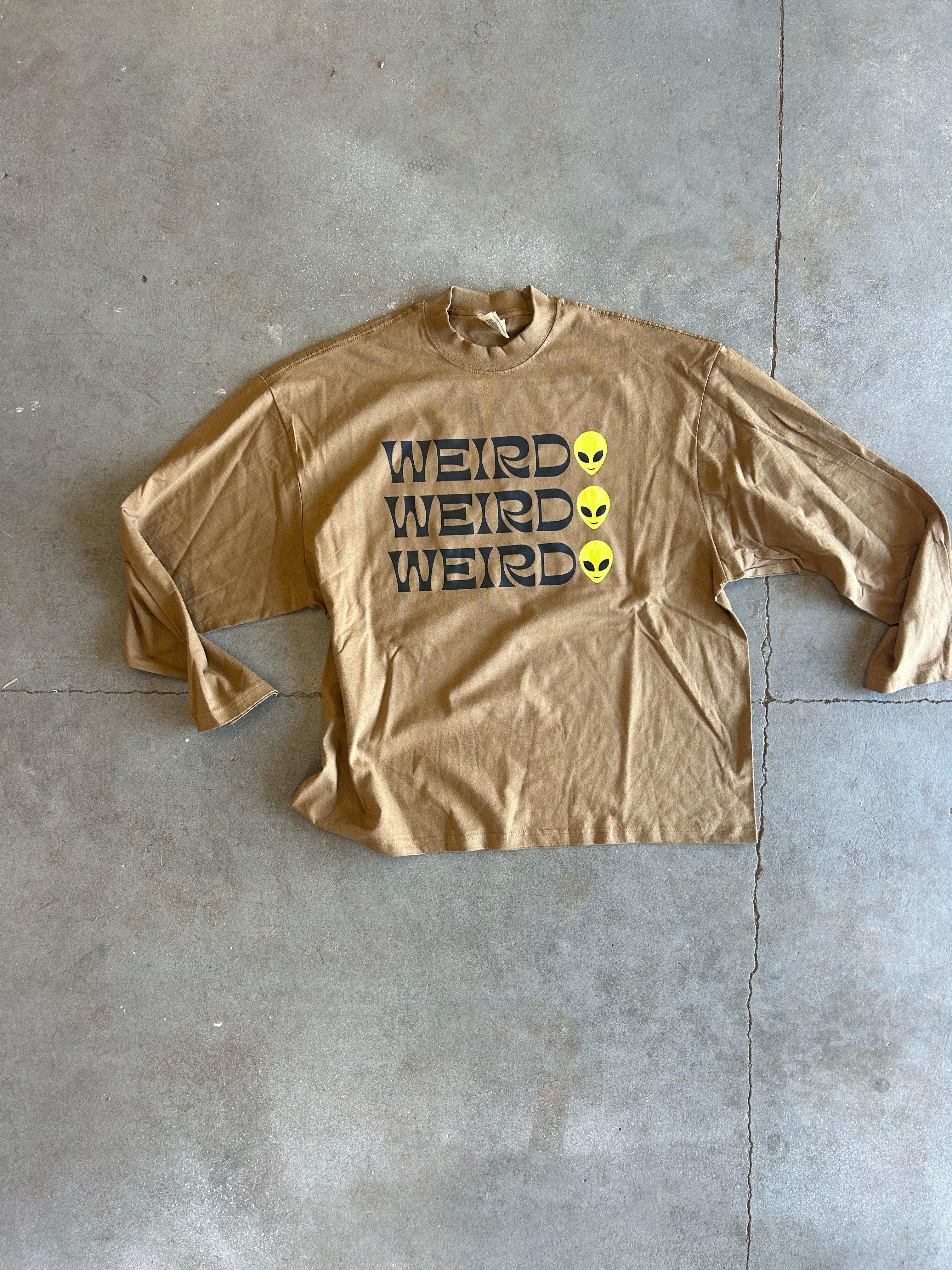 A tan long-sleeve t-shirt with 'WEIRD' printed vertically in black next to three yellow alien heads, laid flat on a gray concrete floor.