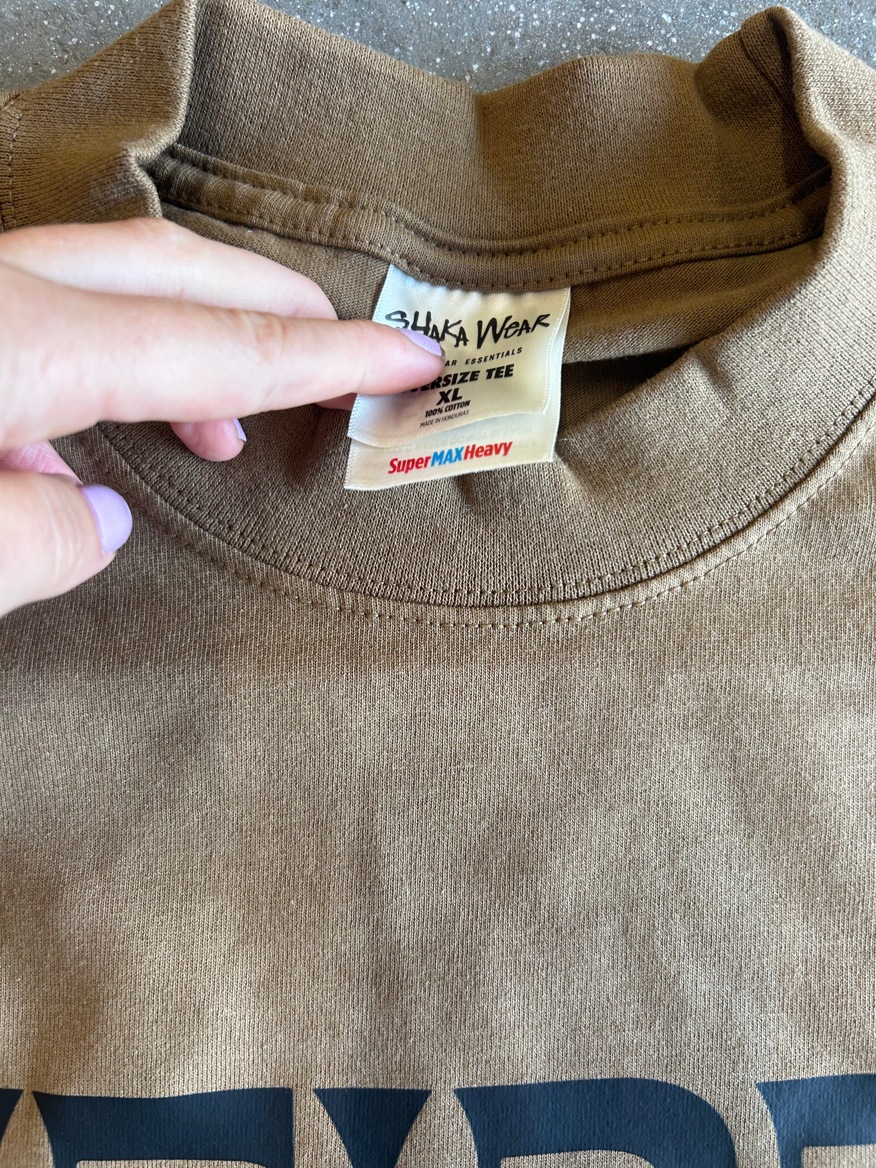 A close-up of a brown crewneck t-shirt showing the inner neck label which reads 'SHARKS WEAR' and 'Super MAX Heavy'. A hand is pointing at the label.