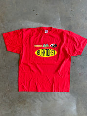 A red short-sleeve t-shirt laid flat on a gray concrete floor, featuring a 'Burntoast' oval logo in yellow and black on the chest, along with smaller sponsor graphics above it.