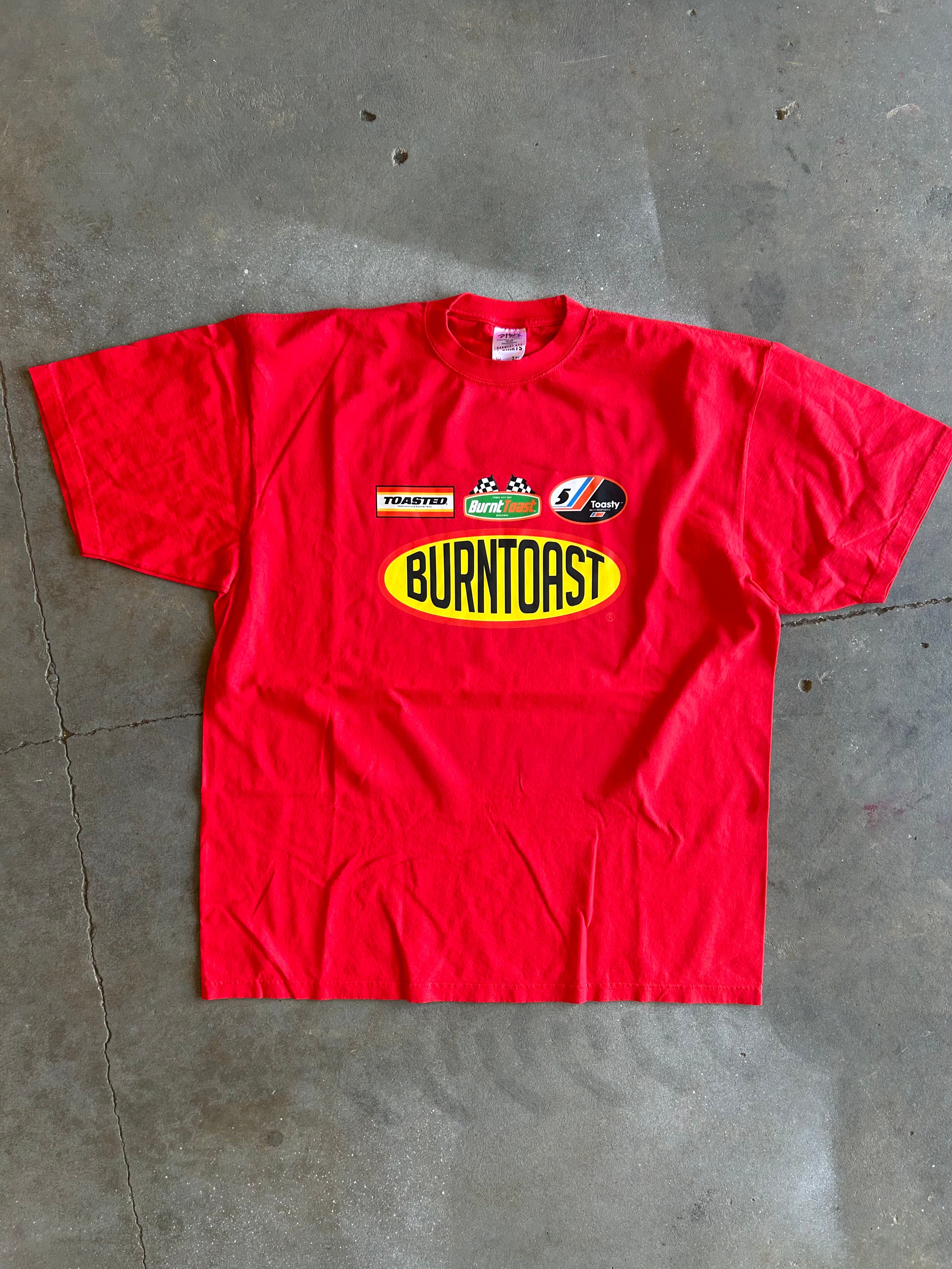 A red short-sleeve t-shirt laid flat on a gray concrete floor, featuring a 'Burntoast' oval logo in yellow and black on the chest, along with smaller sponsor graphics above it.