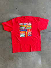 A red short-sleeve t-shirt laid flat on a gray concrete floor, featuring a large graphic print on the back with various colorful racing sponsor logos.